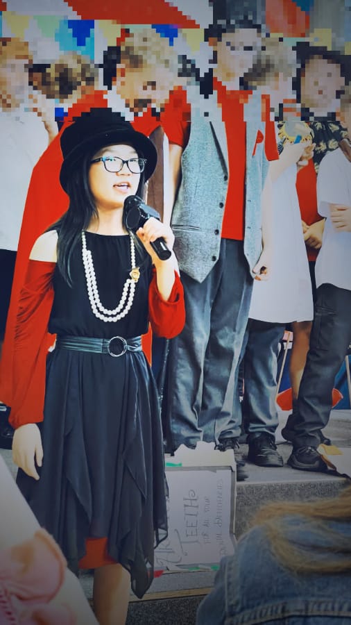 当天发光发亮的主持人 (一位身穿黑色连衣裙和红袖的女孩在舞台上用麦克风说话，背景是模糊的人群)