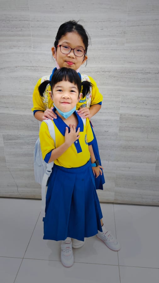 疯猴子一直都表现得比姐姐外向 (两名穿着黄色蓝边校服的女生在浅色木质墙前合影，较高的女孩搂着较矮女孩的肩膀)