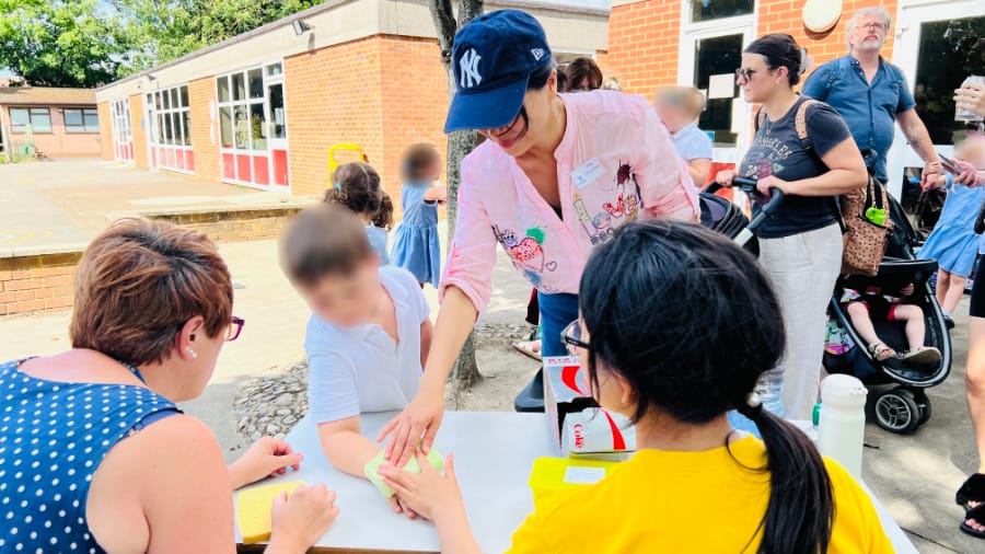 万圣小学的夏季嘉年华 (一位穿粉色上衣和蓝色棒球帽的女士在桌前与一名儿童互动，背景为学校户外活动场地)