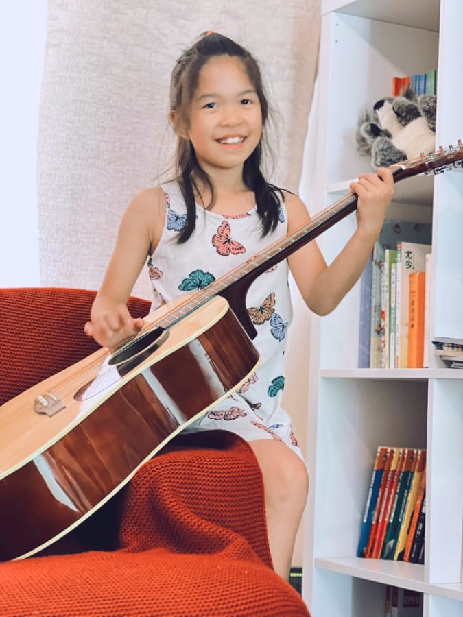 二手吉他带来的意外乐趣 二手吉他带来的意外乐趣 (一名身穿彩色蝴蝶图案白裙的小女孩坐在红椅子上,手持吉他,背景为装满书的白色书架)