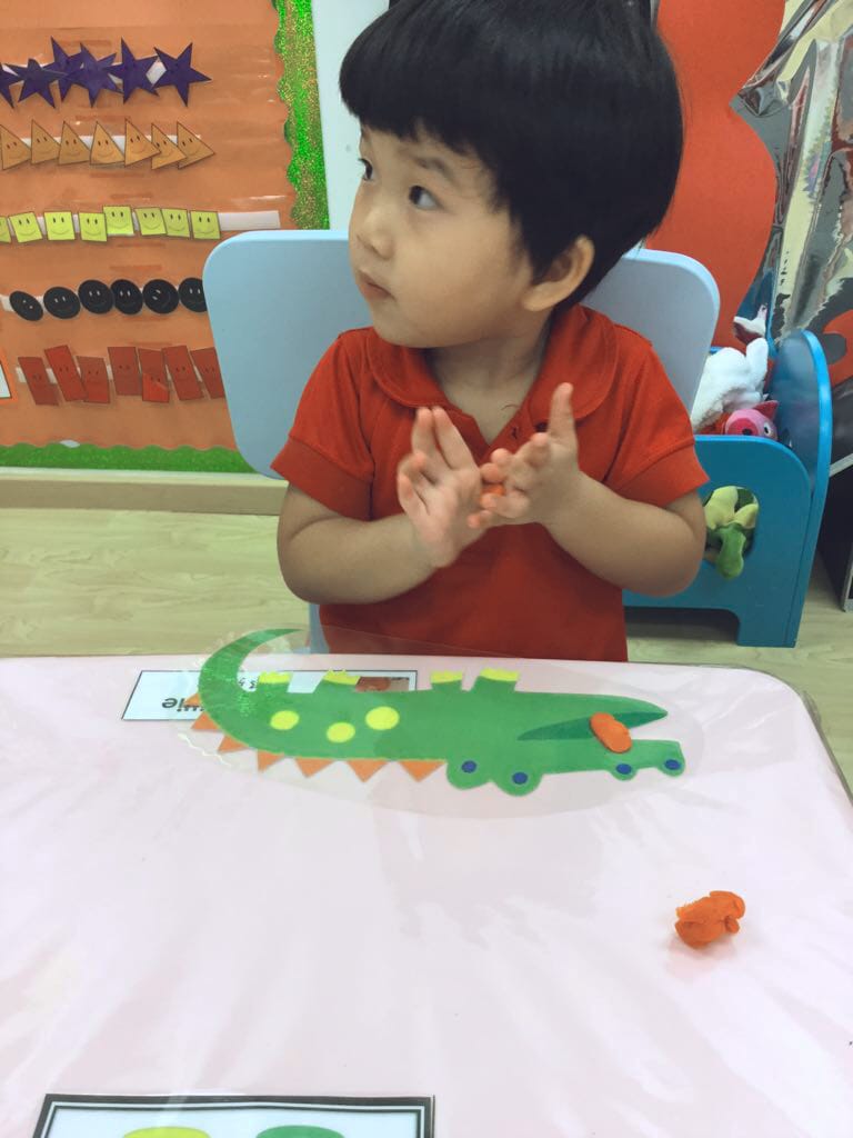 杯杯面在第二所学校的稚嫩样 (幼儿园教室内，一位红衣幼童在粉桌旁玩鳄鱼贴纸和橙色彩泥，背景有蓝椅子与彩色教学挂图)